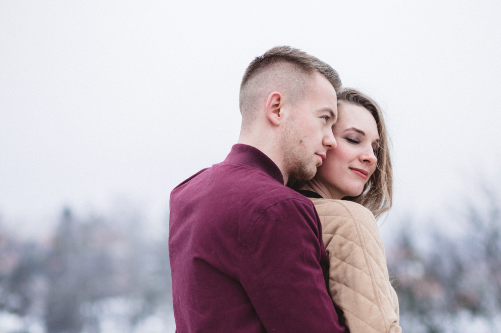 En ung mand står bag en ung kvinde og giver hende et kram. Det ser ud til at det er koldt i vejret. Kvinden smiler med lukkede øjne.